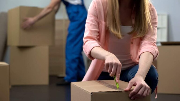 A person wrapping a stack of dishes in bubble wrap at a kitchen table with moving boxes nearby.